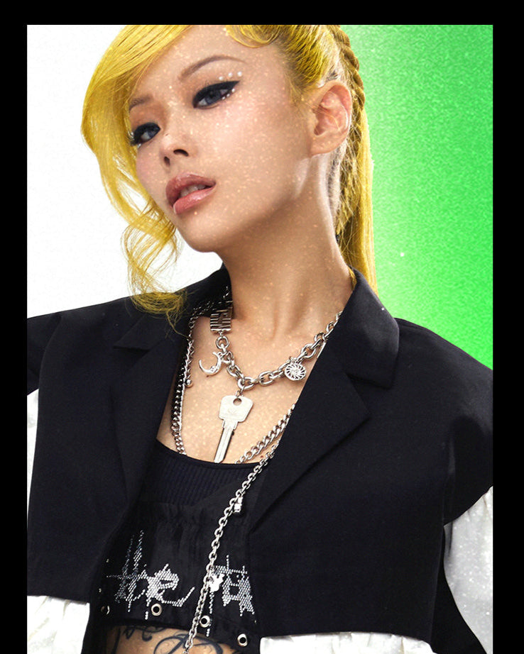 Person with blonde hair wearing a black outfit and multiple necklaces against a green and white background