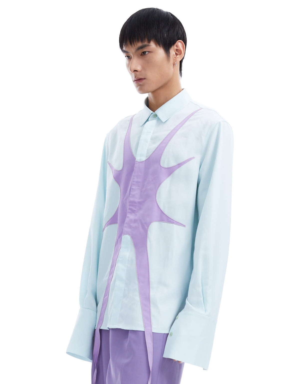 Person wearing a light blue shirt with a purple abstract design and purple pants on a white background