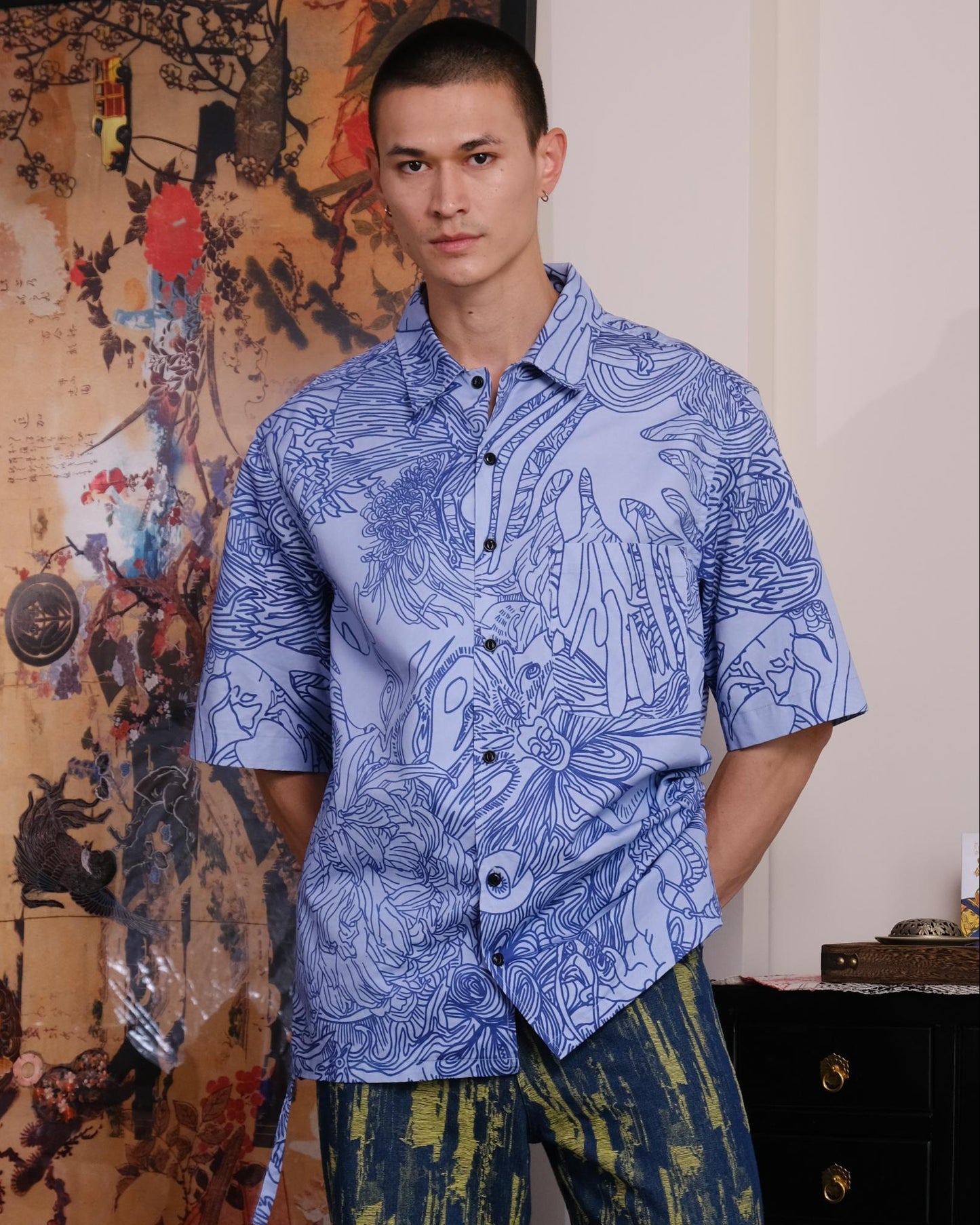 Man wearing a blue patterned shirt and green pants standing in a room with artistic decorations.