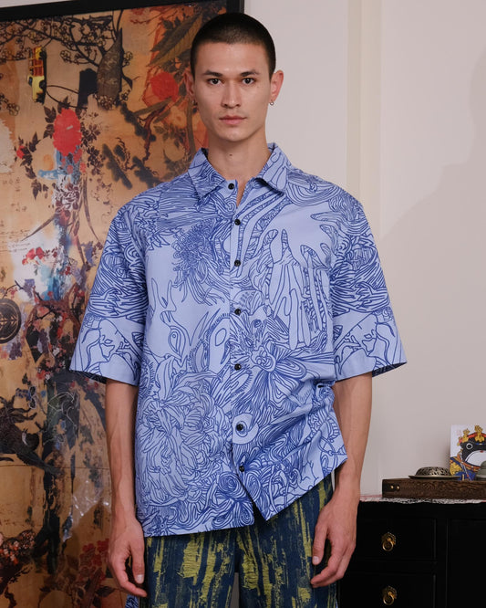 Man wearing a blue patterned shirt and green pants standing in a room with decorative elements.