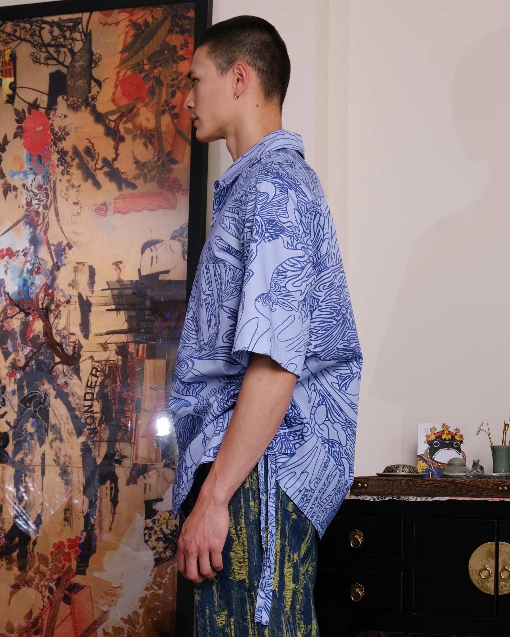 Person wearing a blue patterned shirt and green pants standing in a room with decorative elements.