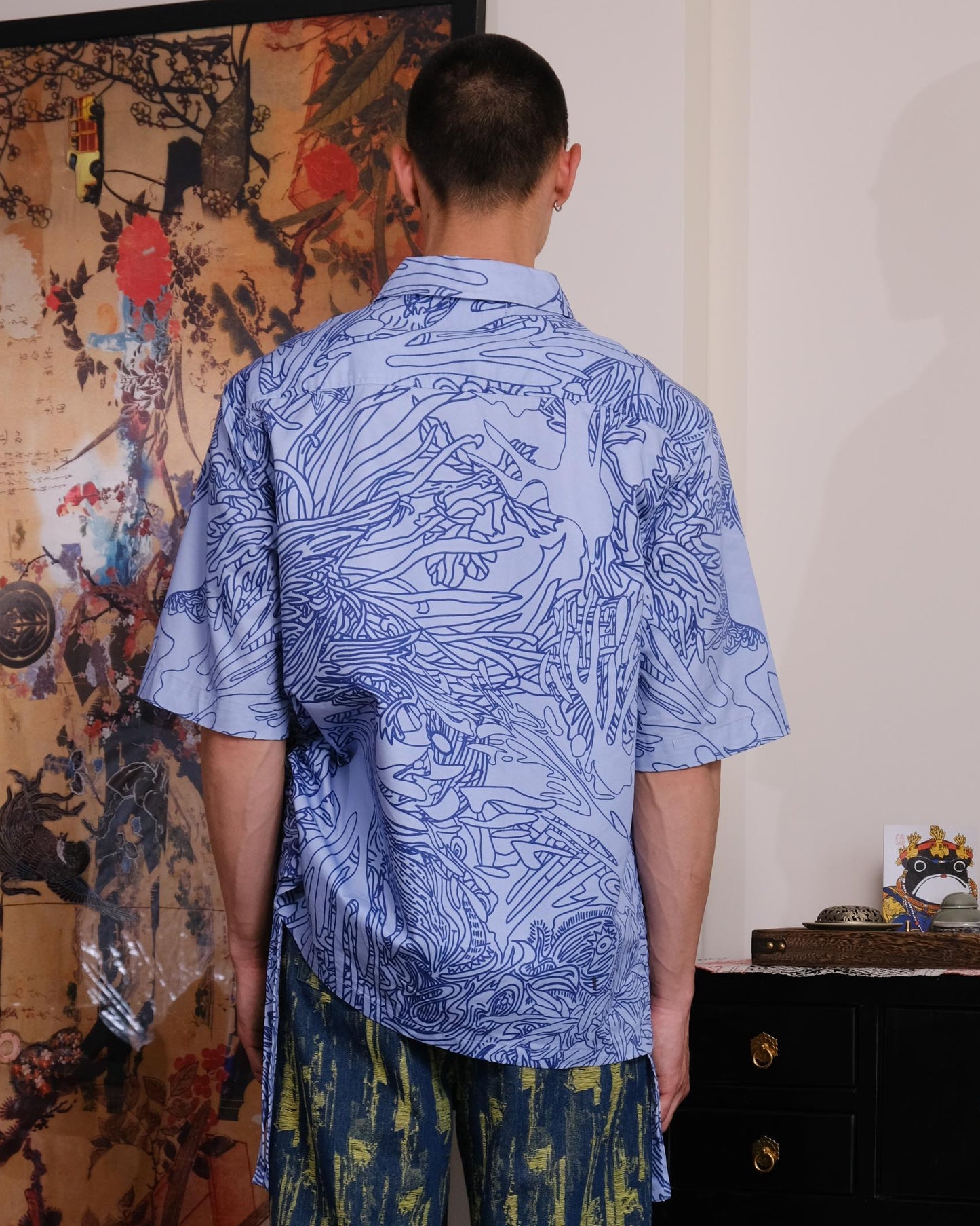 Person wearing a blue patterned shirt and green pants standing in a room with artistic decorations.