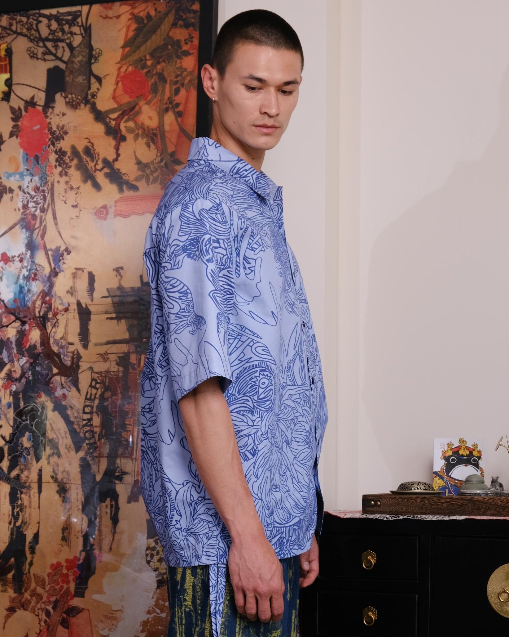 Man in a blue patterned shirt and green pants standing in a room with decorative elements.