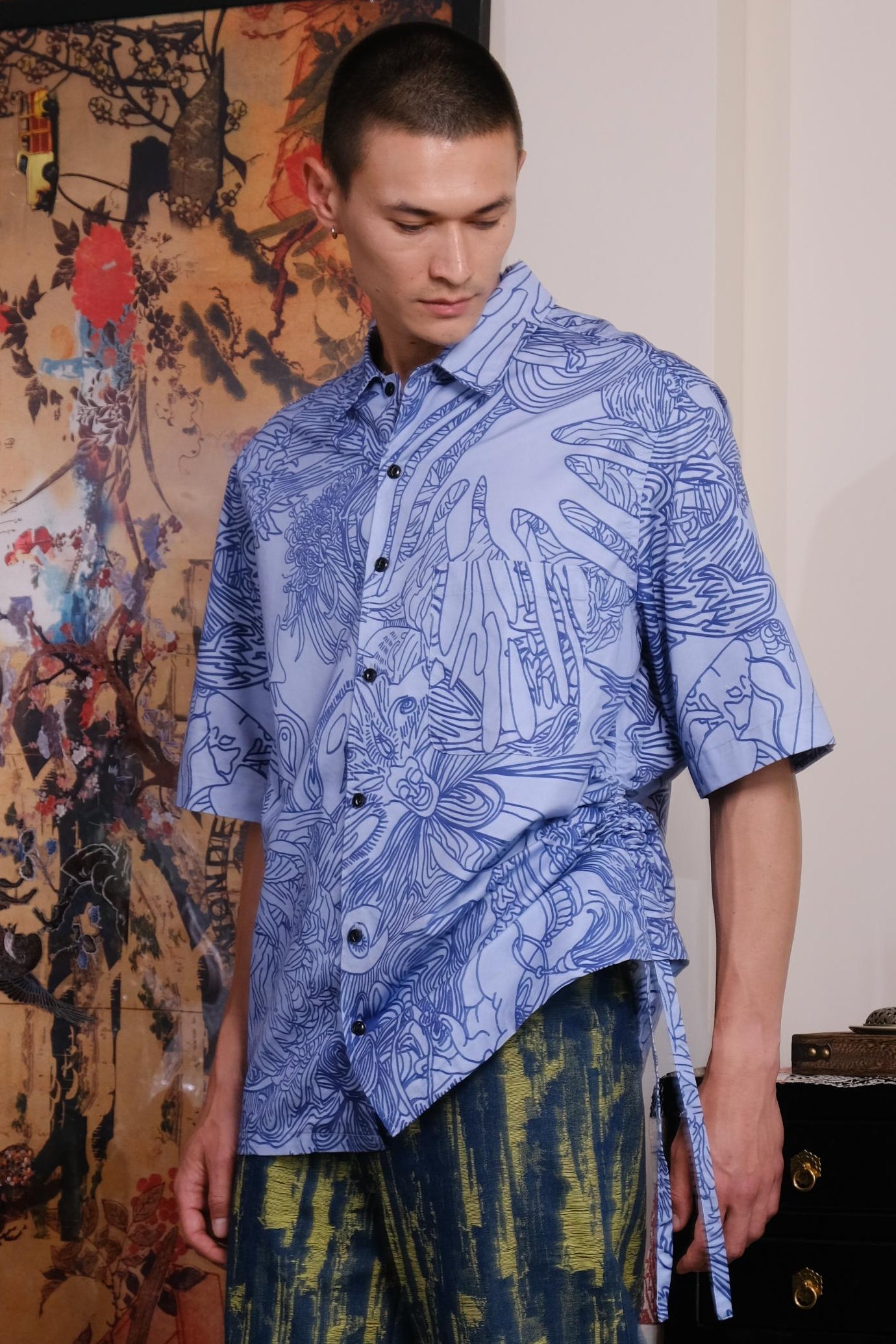 Man wearing a blue patterned shirt and green pants standing in a room with decorative elements.
