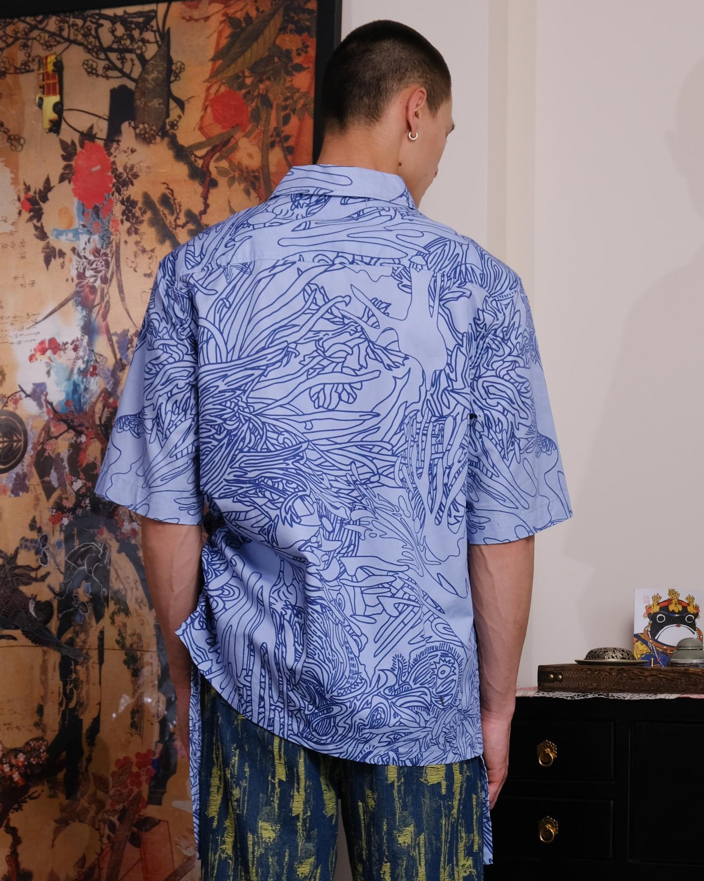 Man wearing a blue patterned shirt and green pants standing in a room with decorative elements.