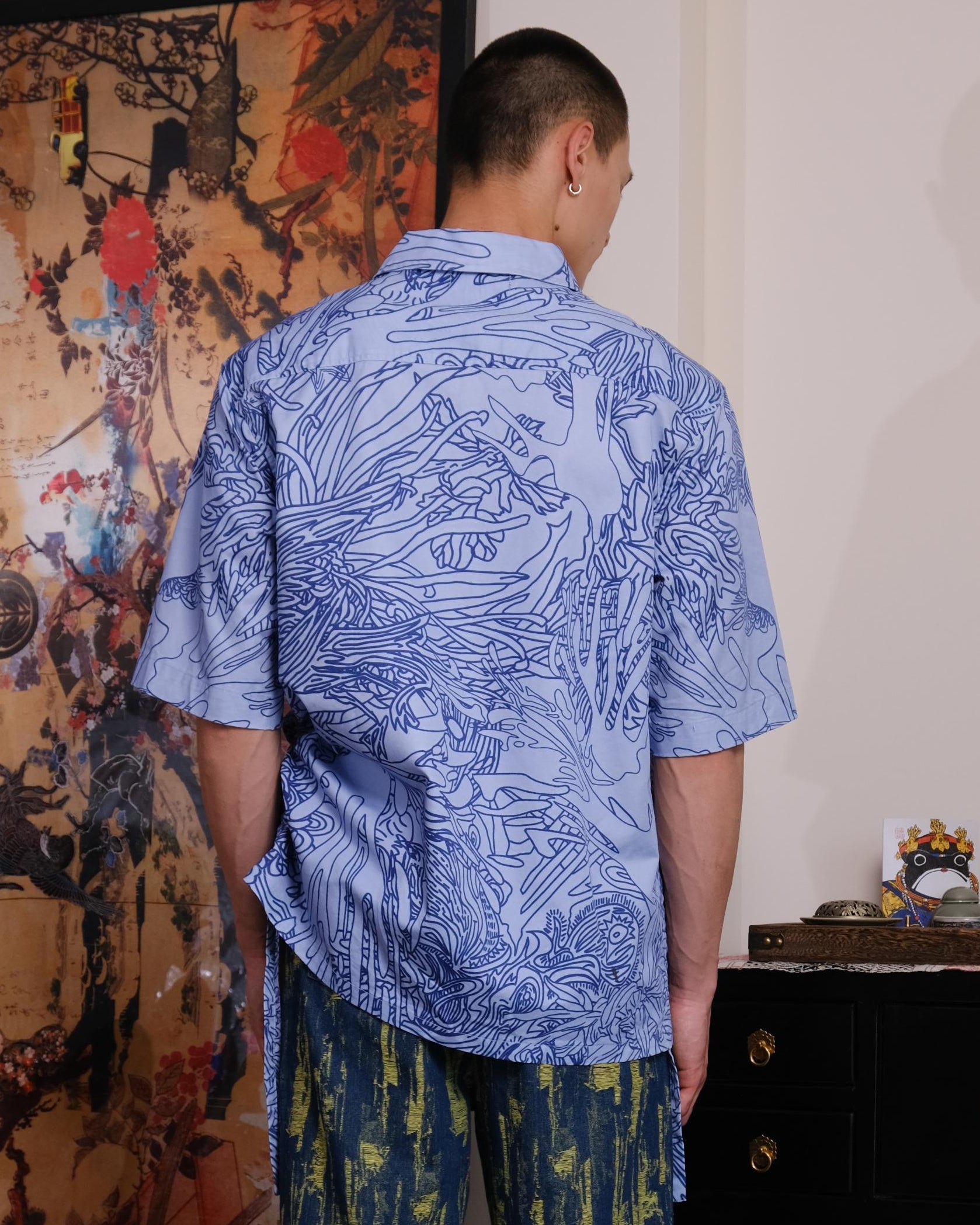 Man wearing a blue patterned shirt and green pants standing in a room with decorative elements.